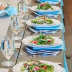 Plates full of colorful salads on a long wooden table with light blue napkins