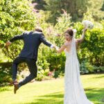 Groom leaping in the air clicking his heels together while bride stands next to him holding his hand and smiling over her shoulder at the camera