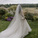 Bride with a veil and a long train with her back to the camera, walking across a grassy lawn towards flowers and a small lake in the background