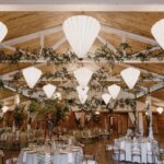 Large dining room with round tables, seating, and place sittings for dinner guests with large inverted pyramid lights and ivy in the rafters