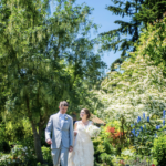 A couple, groom in a grey suit and bride in a sleeveless white dress and a white bouqet walking along a grassy garden with trees and flowers