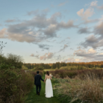 Bride and groom hand in hand walking through tall grass under a lightly clouded sky