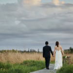 Bride and groom hand in hand walking down a boardwalk through tall grass