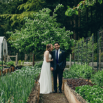 Bride kissing her groom on the cheek as the groom smiles at the camera in hte middle of raised garden beds