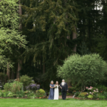 Distant shot of a man with a long white beard and an older woman in a steel blue dress walking arm-in-arm with the bride between the two of them, emerging from a tree-lined path onto a grassy lawn