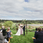 Bride and groom standing hand in hand in front of their officiant with the audience seated in the foreground and a small lake in the background