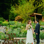 Bride and groom hand in hand looking at each other in a large flower garden with a bench behind them