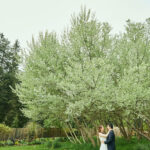 Bride and groom hugging each other in front of a large copse of trees blooming with white flowers