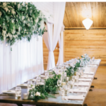 Long table with place settings on both sides, white flowers and greenery up the middle of the table, and curtains and more greenery and white flowers hanging to the left of the table