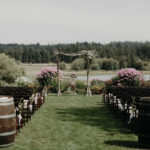 Two wine barrels behind rows of wooden chairs overlooking a birch wedding arch in front of a small lake