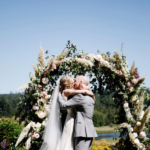 Man in a light grey suit and woman in a white dress kissing with arms wrapped around one another beneath an arch of large white flowers and ferns