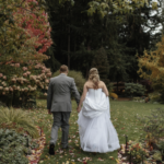 Woman in a large skirt white dress and man in a grey suit walking hand in hand away from the camera across a grassy lawn with fall leaves on the trees and scattered around them