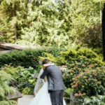 Man in a grey suit and woman in a white dress, man's back to the camera, kissing in the middle of tall bushes of greenery and flowers