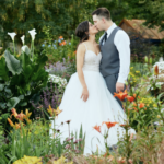 Man in grey vest and woman in white dress with arms to their sides and kissing with flowers in the foreground and to the left of the couple