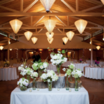 Rectangular table of white flower bouqets in front of a reception hall full of round tables and inverted pyramid lights
