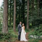 Man in grey suit and woman in white standing side by side on a forest dirt path with tall skinny trees around them