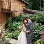 Man in a grey suit kissing the side of a woman in a white dress's face as she looks at the camera, in front of flowers and bushes with the brown wood barn in the background