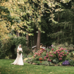 Bride holding a bouqet and walking across a grassy lawn with flowers in the background and trees behind her